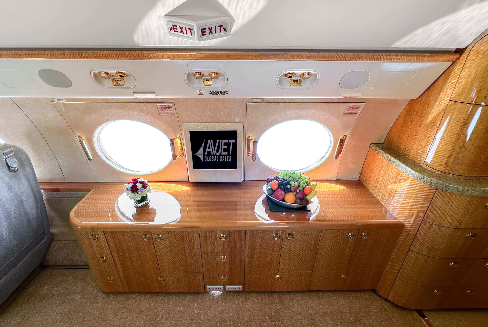 The aft cabin credenza of this 2010 Gulfstream G550 showcases exquisite craftsmanship with high-gloss wood veneer, polished gold hardware, and elegant storage cabinetry. Positioned beneath two large oval windows, the space is beautifully accented with natural light, creating a refined area ideal for serving, storage, or display during flight.
