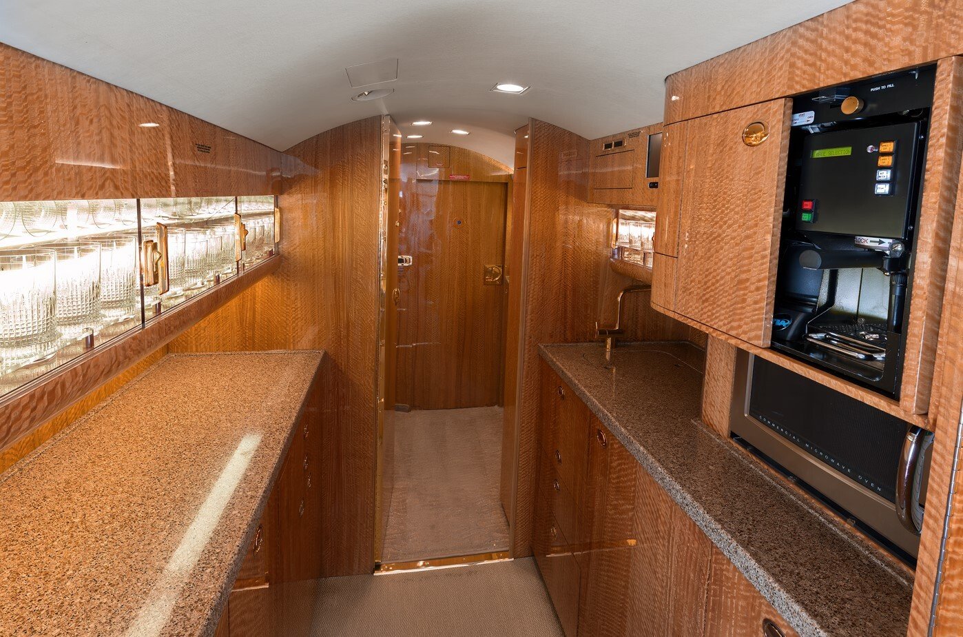 The aft galley of this 2010 Gulfstream G550 is designed for both functionality and elegance, featuring dual granite countertops, fine wood cabinetry, and illuminated crystal glassware displays. Equipped with premium appliances, ample storage, and gold-accented fixtures, it provides a refined space for meal preparation and in-flight service.