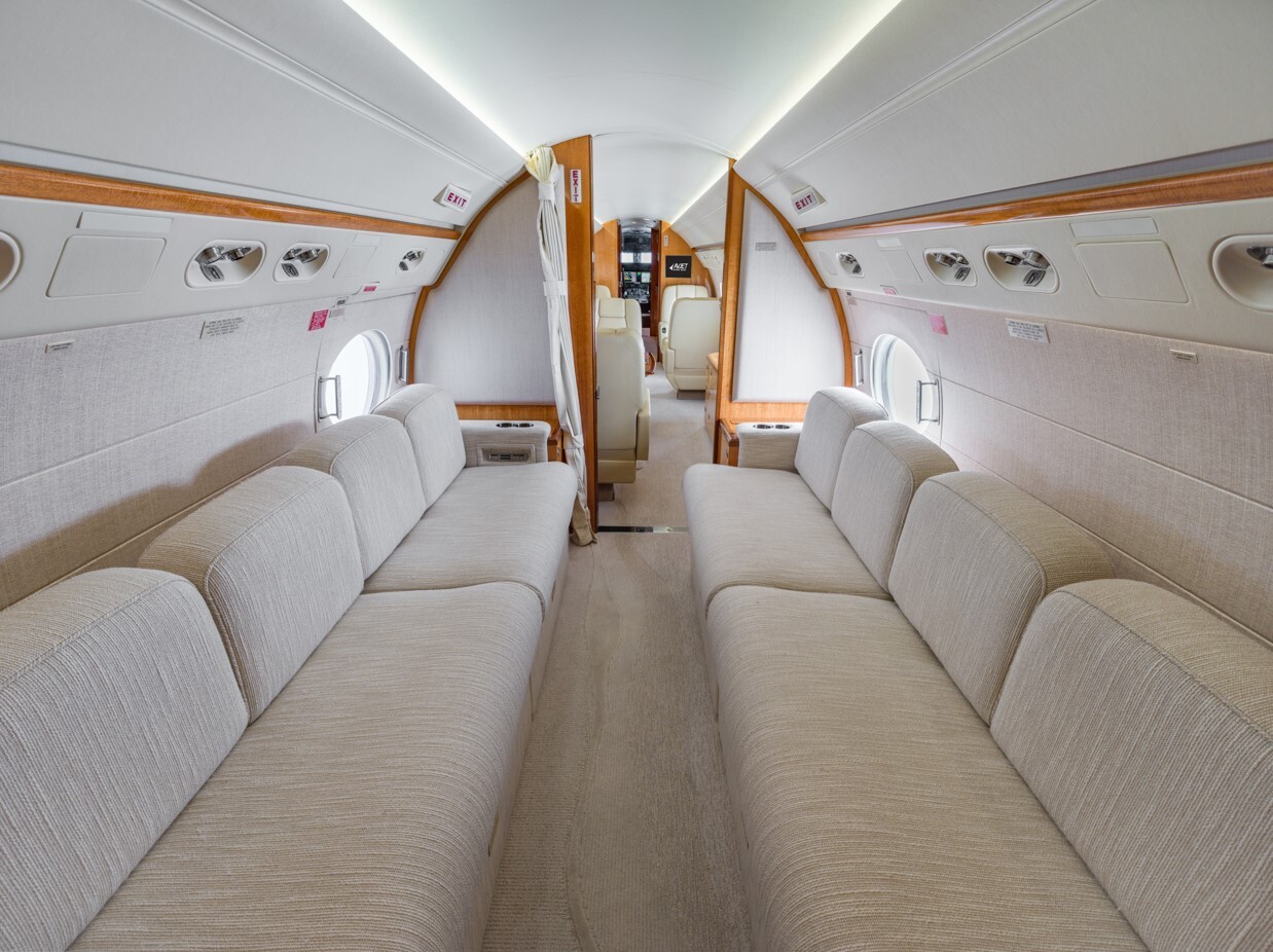Aft cabin of a Gulfstream 550, view looking forward, featuring dual opposing cream divans, panoramic oval windows, and direct access to the mid cabin—offering a spacious and versatile environment for relaxation, entertainment, or crew rest on long-range missions.