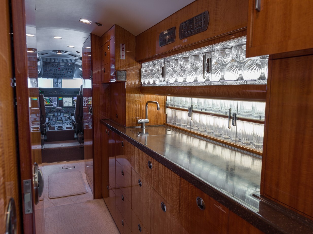 Forward galley of a Gulfstream G550, showcasing high-gloss wood cabinetry, integrated sink, glassware storage with backlit crystal display, and direct view into the cockpit—designed for efficient in-flight catering and elegant service.