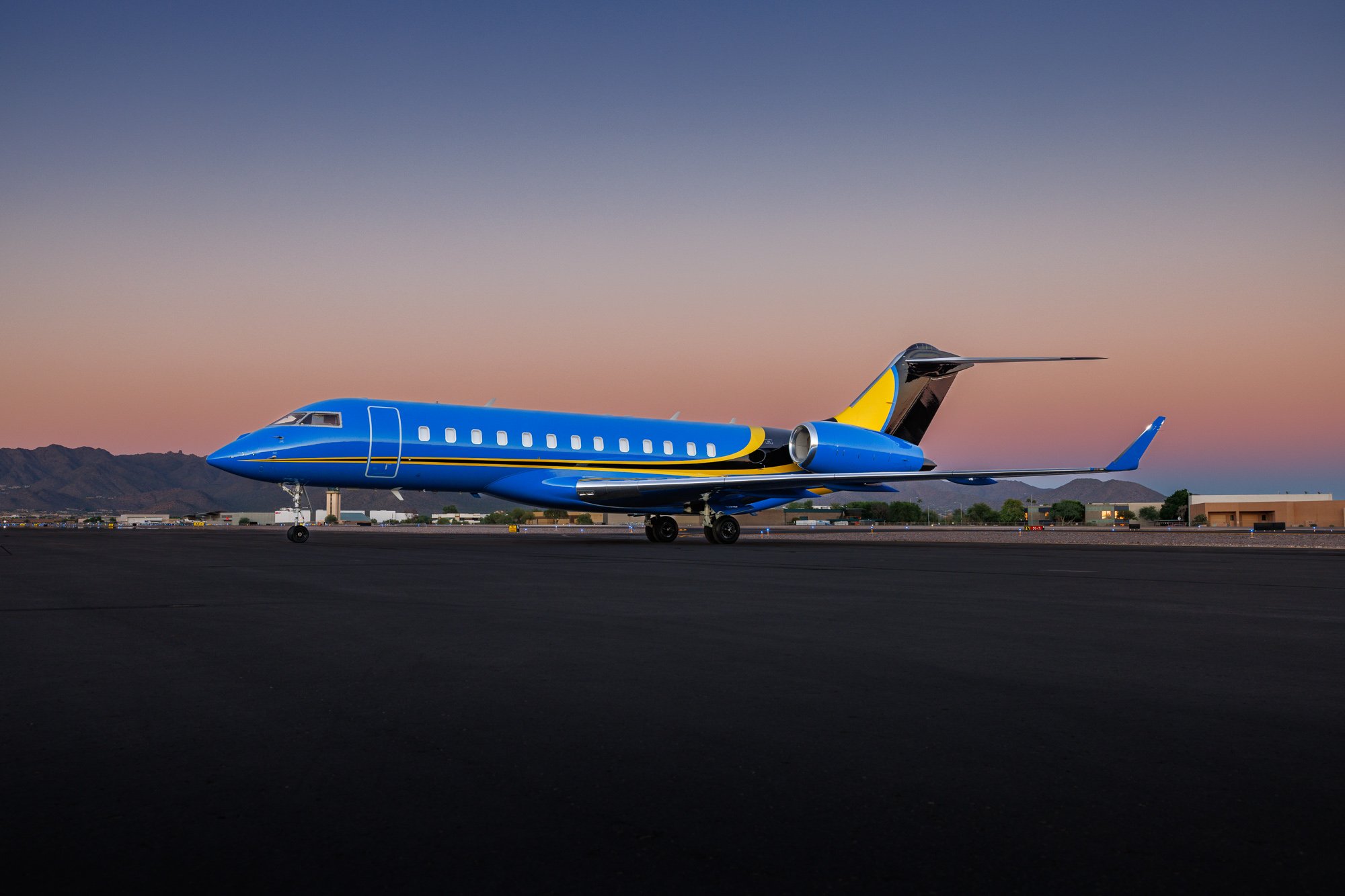 Bombardier Global private jet in striking blue and yellow livery parked on the runway at sunset, showcasing luxury, performance, and long-range capability — photographed against a mountain backdrop and gradient sky.