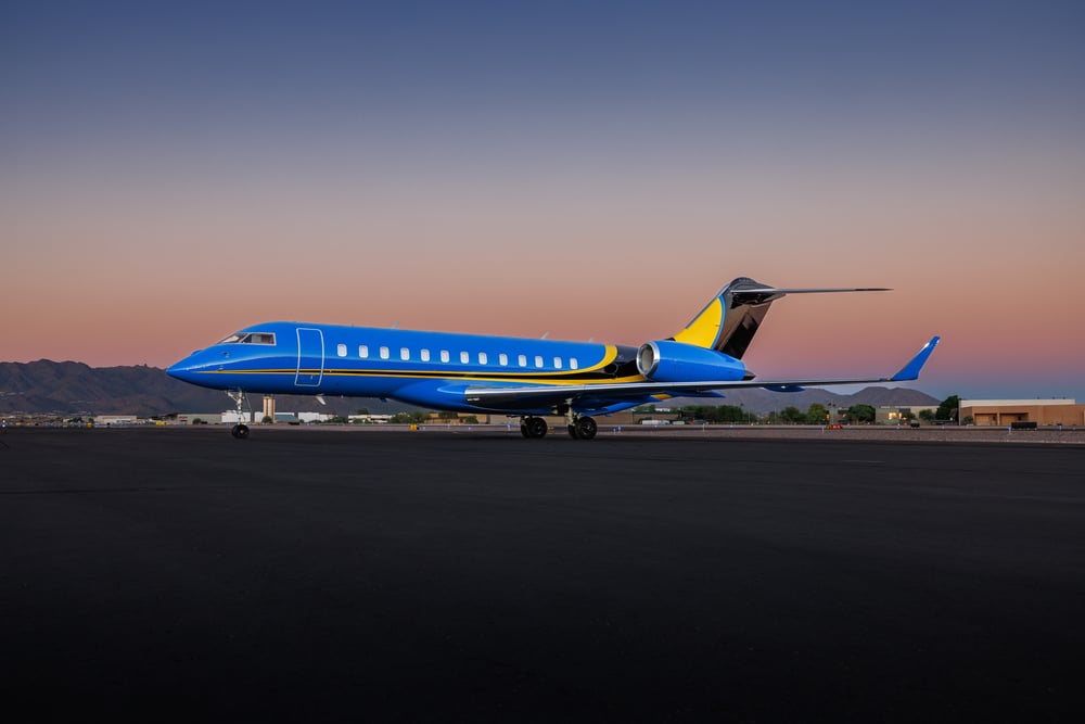 Bombardier Global private jet in striking blue and yellow livery parked on the runway at sunset, showcasing luxury, performance, and long-range capability — photographed against a mountain backdrop and gradient sky.