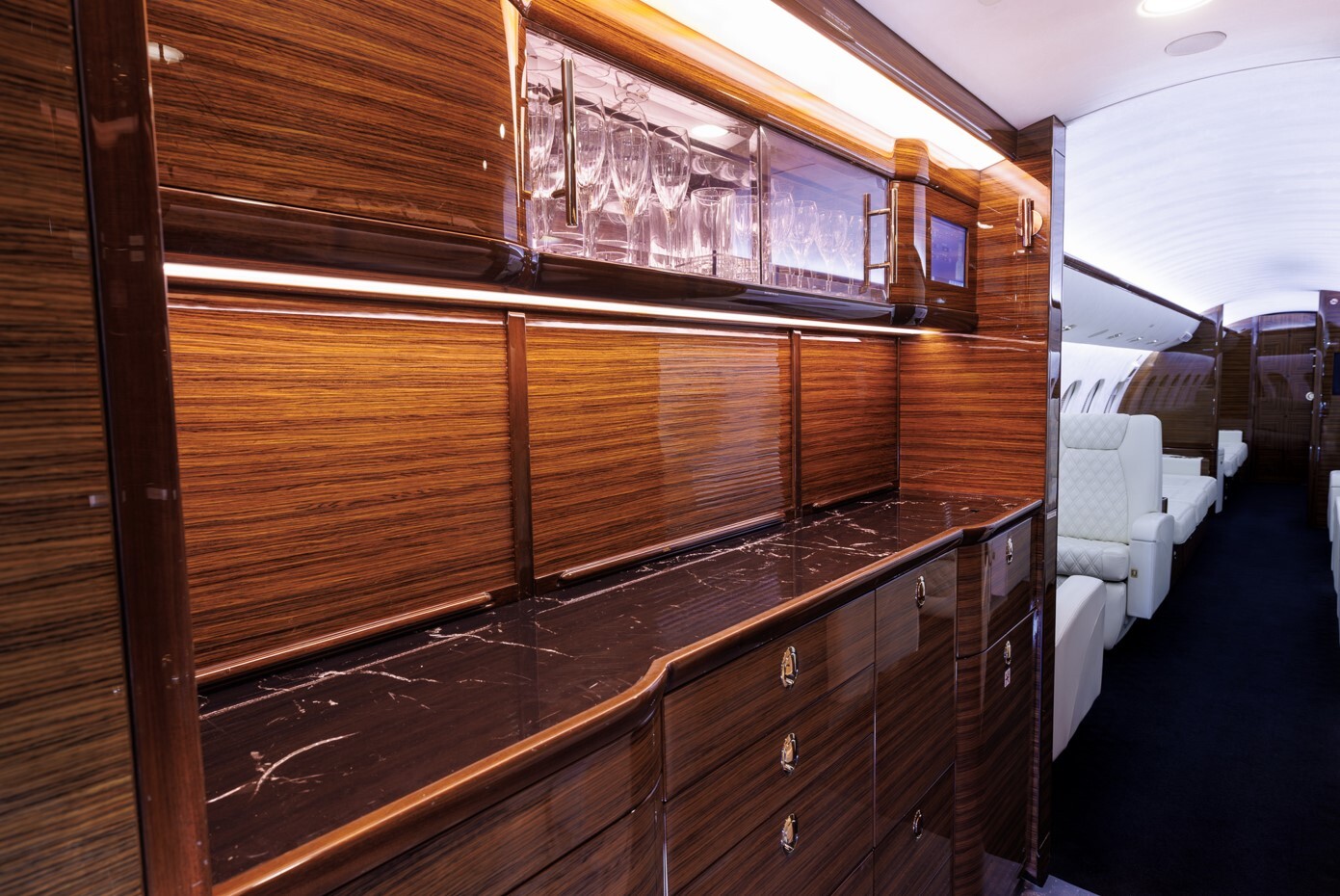 Forward galley of a Bombardier Global private jet featuring high-gloss wood cabinetry, black marble countertop, and illuminated glassware storage — designed for elegant in-flight service and convenience.