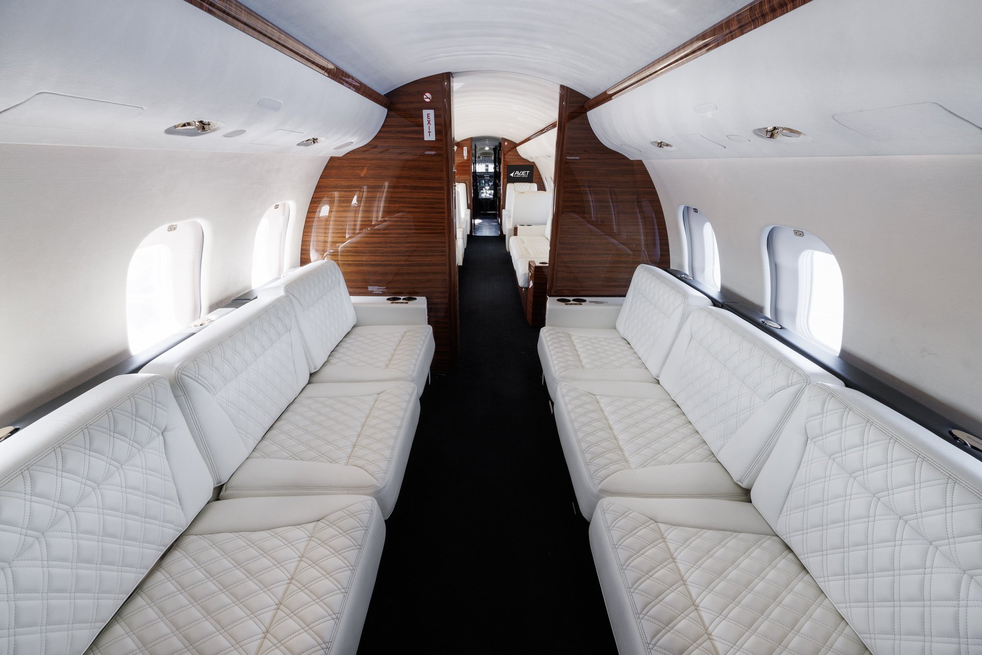 Aft cabin view looking forward inside a Bombardier Global private jet, showcasing dual opposing three-place divans upholstered in white diamond-stitched leather, with rich wood paneling and sleek cabin design for refined in-flight comfort.