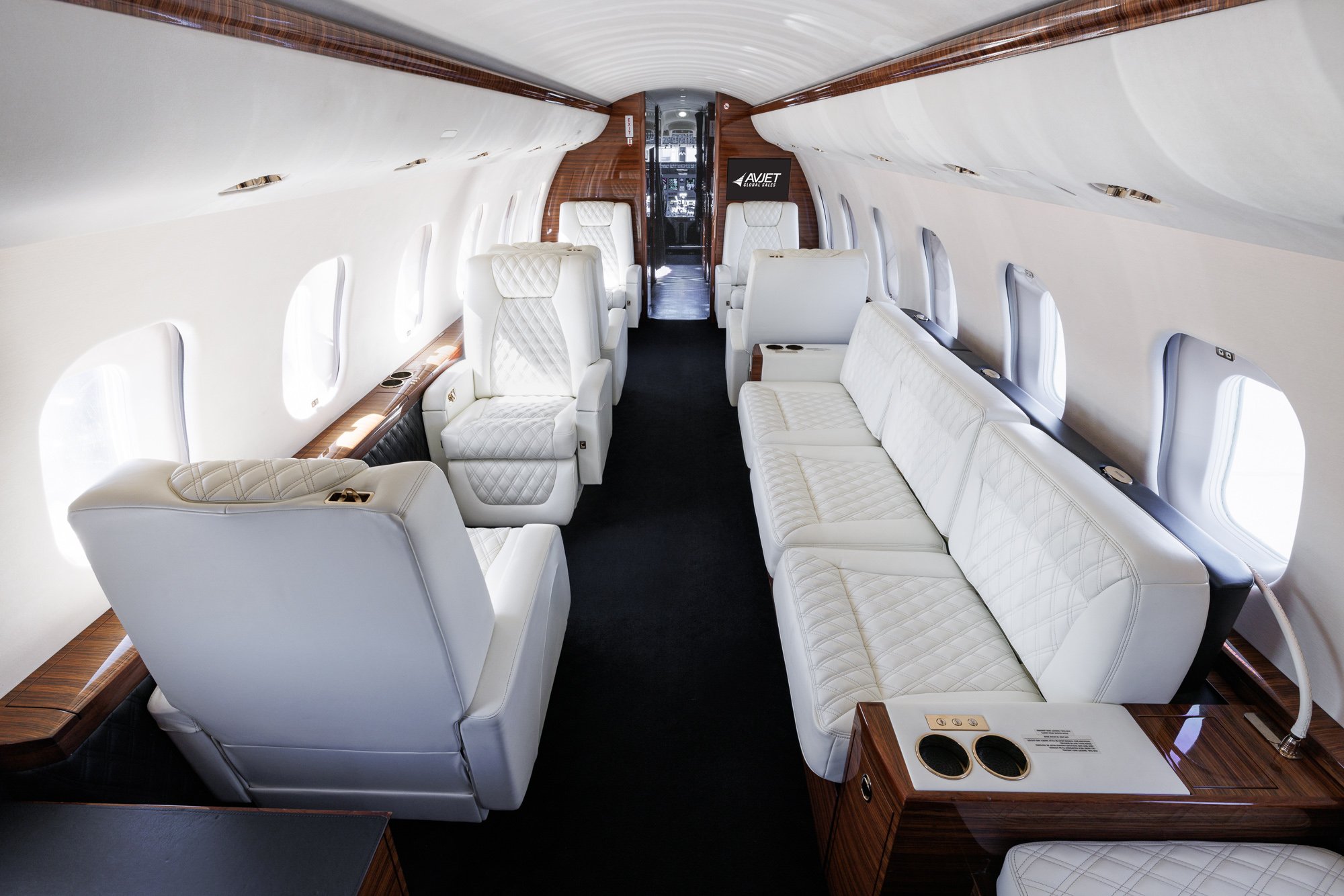 Mid cabin view looking forward inside a Bombardier Global private jet, featuring elegant white diamond-stitched leather seats, a three-place divan, and rich wood accents, offering a seamless transition to the forward lounge and cockpit.