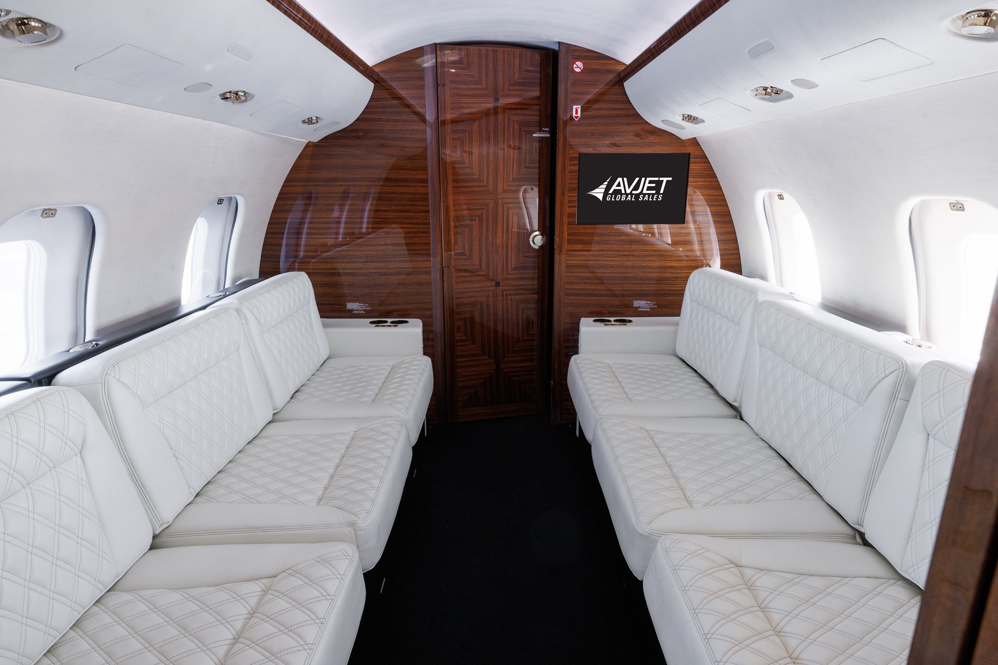Aft cabin view looking aft inside a Bombardier Global private jet, featuring dual three-place divans upholstered in white diamond-stitched leather, accented with high-gloss wood paneling for a refined and spacious lounge environment.
