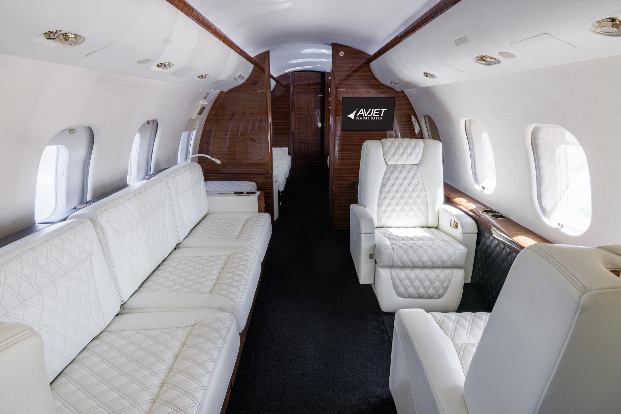 Mid cabin view looking aft inside a Bombardier Global private jet, featuring white diamond-stitched leather club seating and a three-place divan, complemented by rich wood accents and panoramic windows.