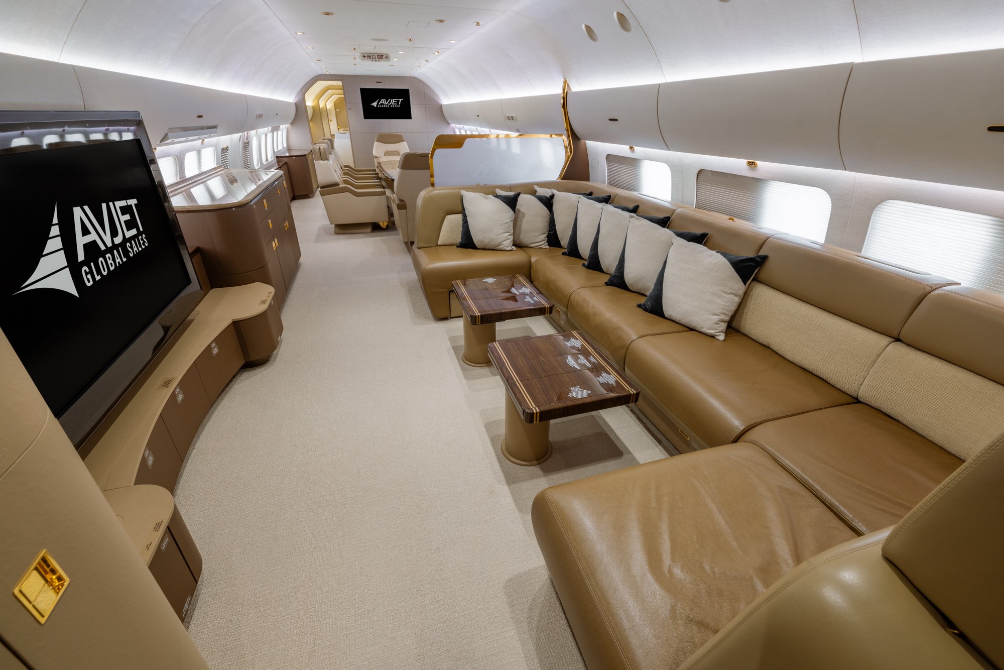 The expansive VIP Lounge of the 2018 Boeing Business Jet 2 (BBJ2) features a luxurious wraparound leather sofa, handcrafted wood tables, and a large entertainment display, creating an inviting space for relaxation, socializing, and in-flight entertainment during long-range journeys.
